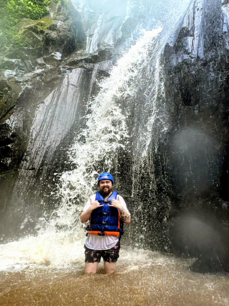 A transgender man stands confidently in a pool of water beneath a massive Indonesian waterfall during a canyoning adventure. He wears a blue safety helmet and life jacket, making shaka hand signs to celebrate the outdoor exploration and travel experience.