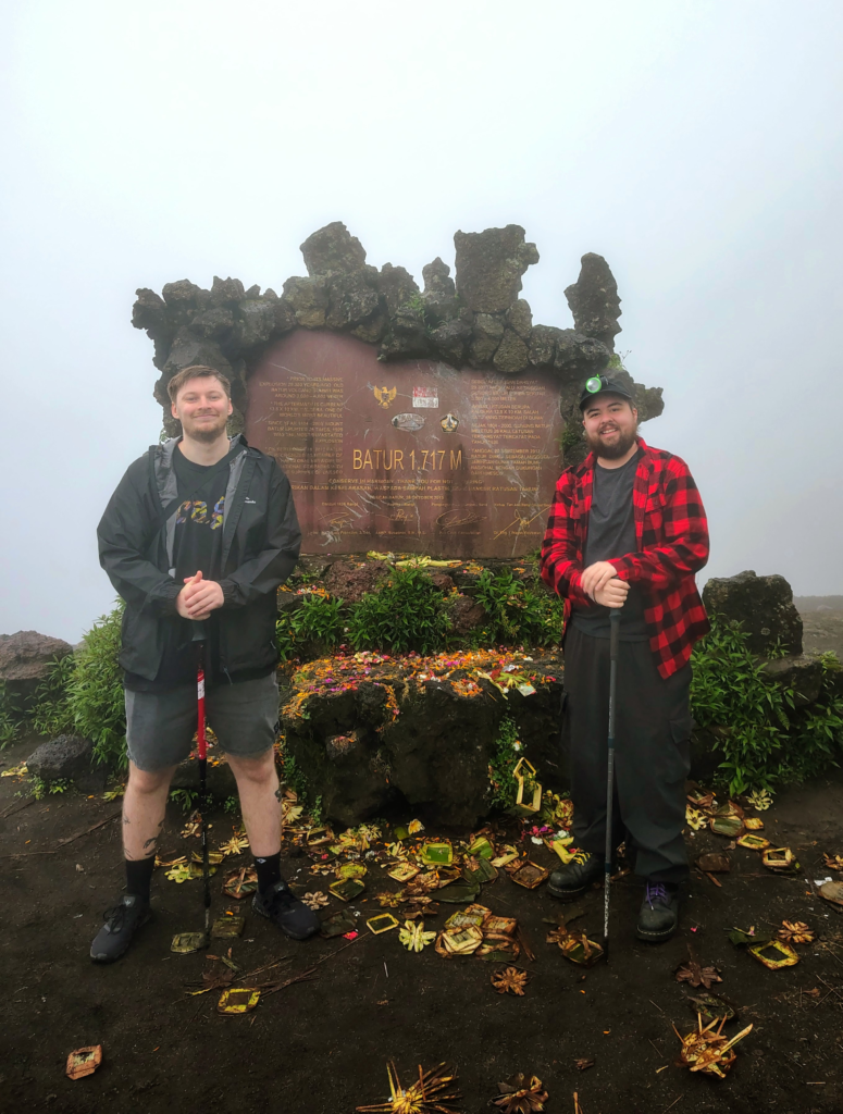 A transgender man and a friend stand proudly at the summit of Mount Batur in Bali, Indonesia, next to a stone marker reading "BATUR 1,717 M." Both hikers hold trekking poles and smile against a backdrop of thick mountain mist, surrounded by traditional Balinese offerings on the volcanic ground.