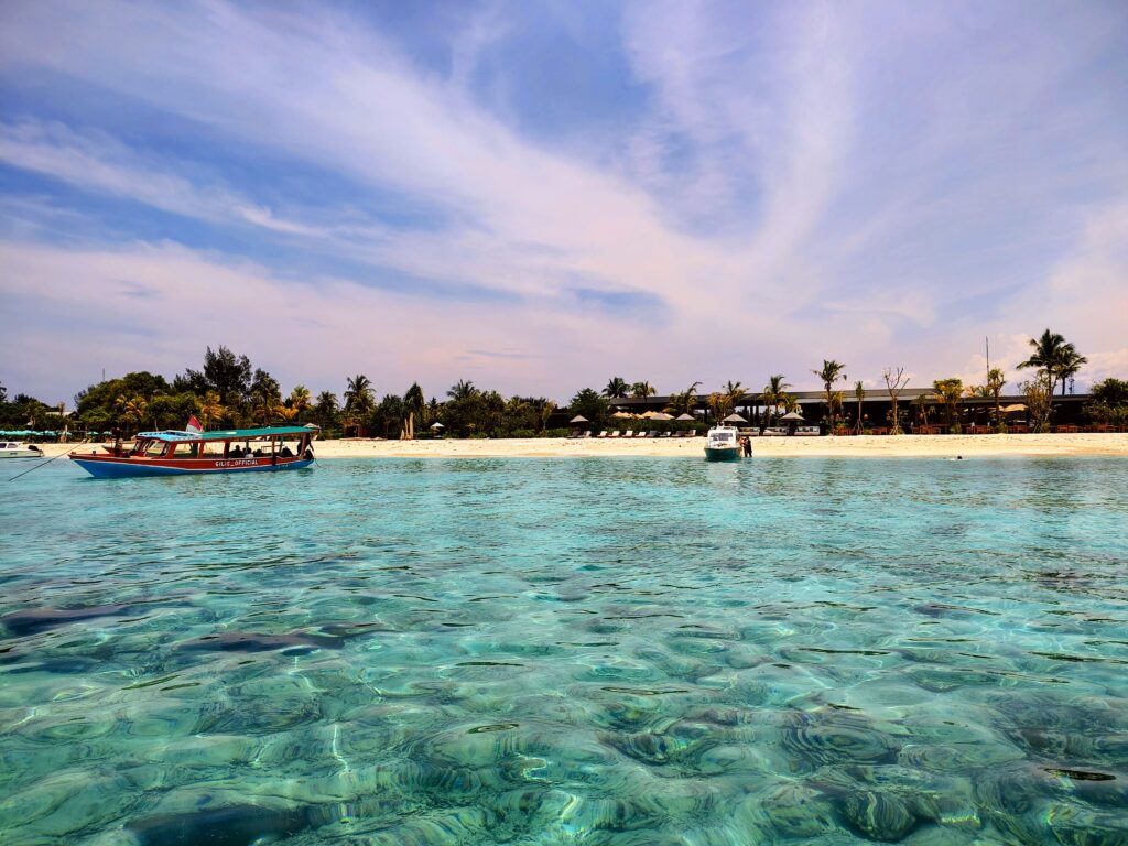 Clear, turquoise water ripples in the foreground of a scenic beach on the Gili Islands, Indonesia. A red and blue traditional boat floats near a white sand shore lined with palm trees and a beachfront resort under a bright, wispy blue sky.