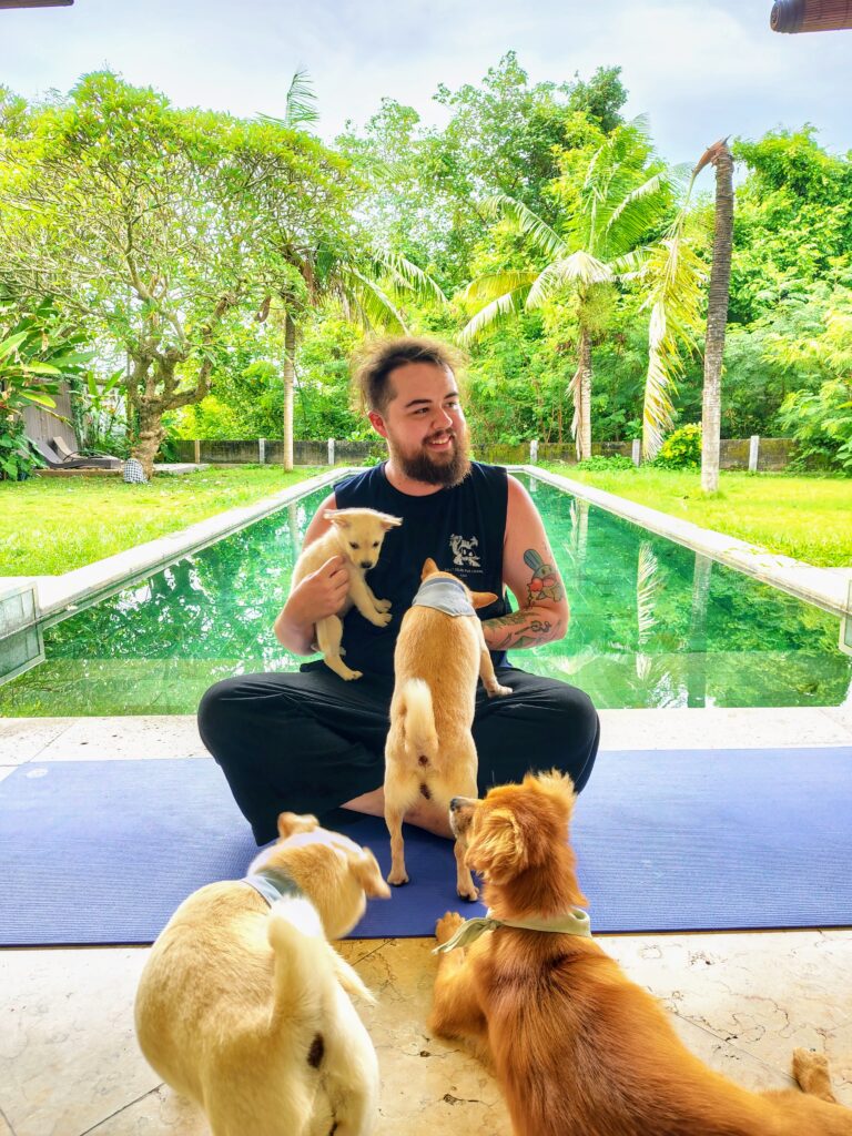 A transgender man sits cross-legged on a blue yoga mat by a turquoise swimming pool in Bali, Indonesia, surrounded by several small, friendly dogs. He smiles as he holds a light-colored puppy, reflecting the peaceful and welcoming atmosphere of his Indonesian travel journey.