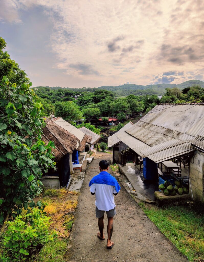 A view from behind a person walking down a sloping concrete path in a hillside village. The person wears a white shirt, grey shorts, and a blue towel draped over their shoulders. On either side of the path are simple structures; one features a pile of green coconuts. In the background, a thick canopy of tropical trees stretches across the hills under a vast sky filled with soft, textured clouds.