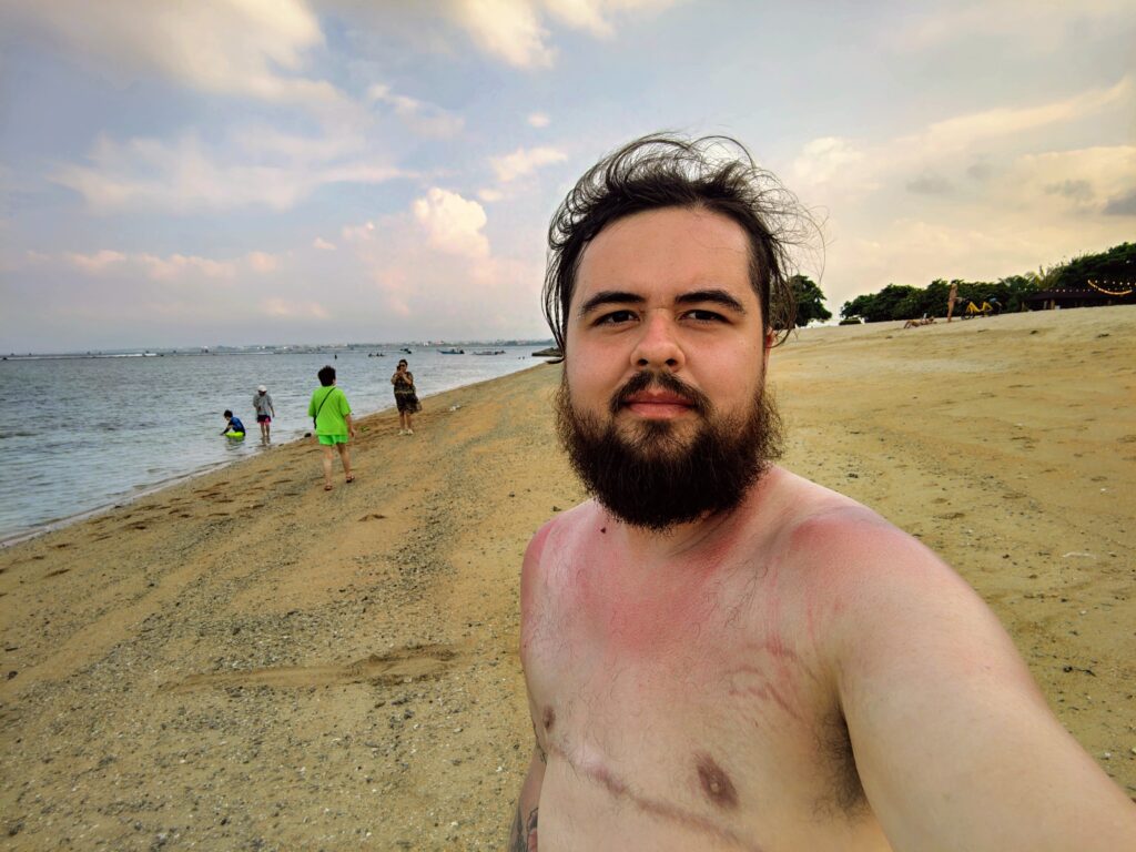 A shirtless transgender man with a beard and visible top surgery scars takes a selfie on a tropical beach in Indonesia. He stands on the sand with the ocean and a soft, cloudy sunset sky behind him, reflecting a moment of body confidence and authenticity during his travels.