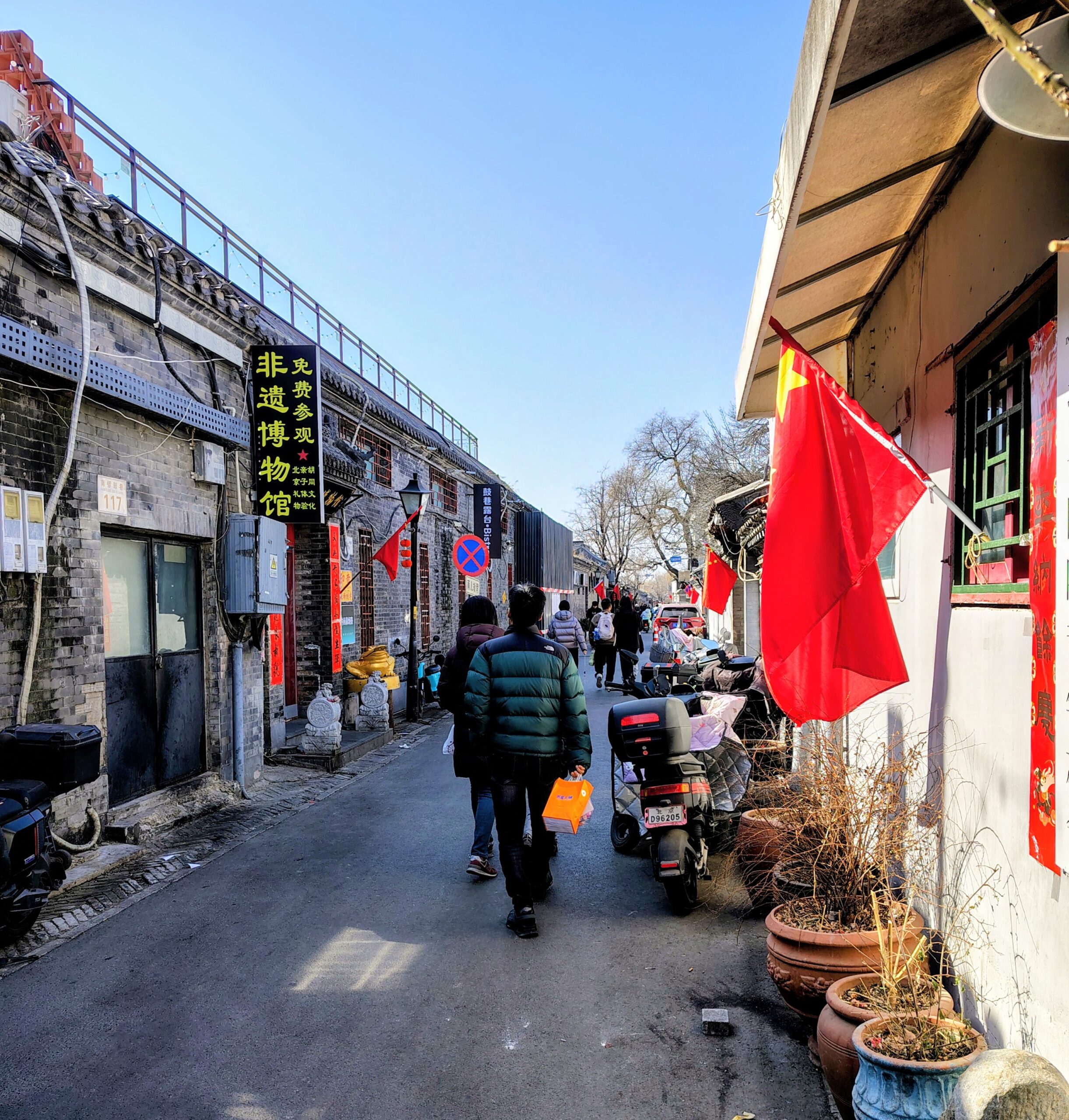 A street-level view of a narrow hutong alley in Beijing, China, lined with grey brick buildings and adorned with vibrant red Chinese flags. Pedestrians walk away from the camera past parked motorbikes and potted plants, illustrating the daily life and atmosphere encountered by a transgender man traveling through China’s historic neighborhoods.