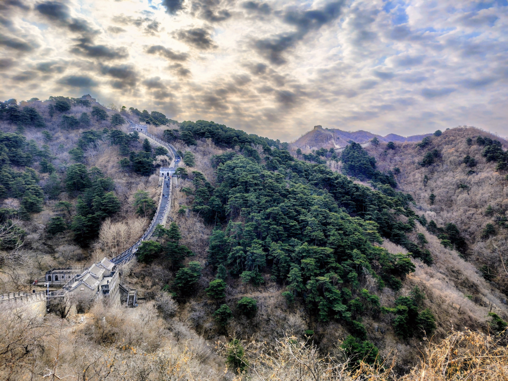 A dramatic, wide-angle photograph of the Great Wall of China, showing its steep stone stairway and multiple watchtowers winding over rugged, heavily forested mountains. The sky is dramatic with heavy, textured grey and white clouds, but patches of bright, radiant sunlight break through. This image of challenging ascent symbolizes the transformative journey of a transgender man traveling in China, combining iconic sights with an empowering narrative of resilience and self-discovery.