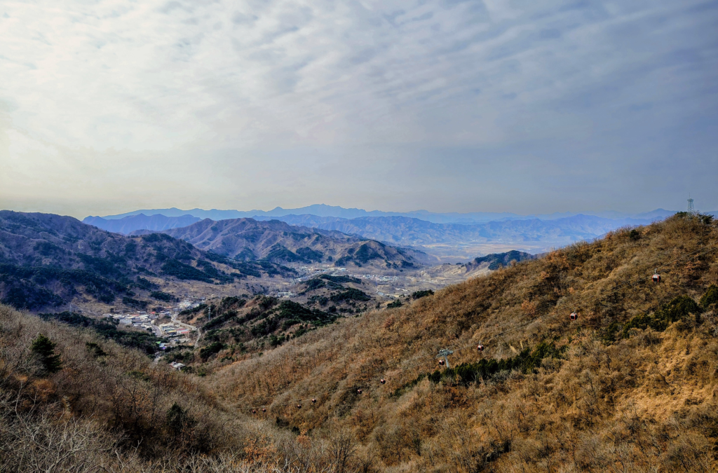 A wide, panoramic view of the vast, rolling mountains surrounding the Great Wall of China under a hazy, patterned sky. Cable cars scale the arid, tree-dotted hillside on the right, overlooking a small village in the valley below; a landscape feature from an LGBTQ+ and trans travel blog.