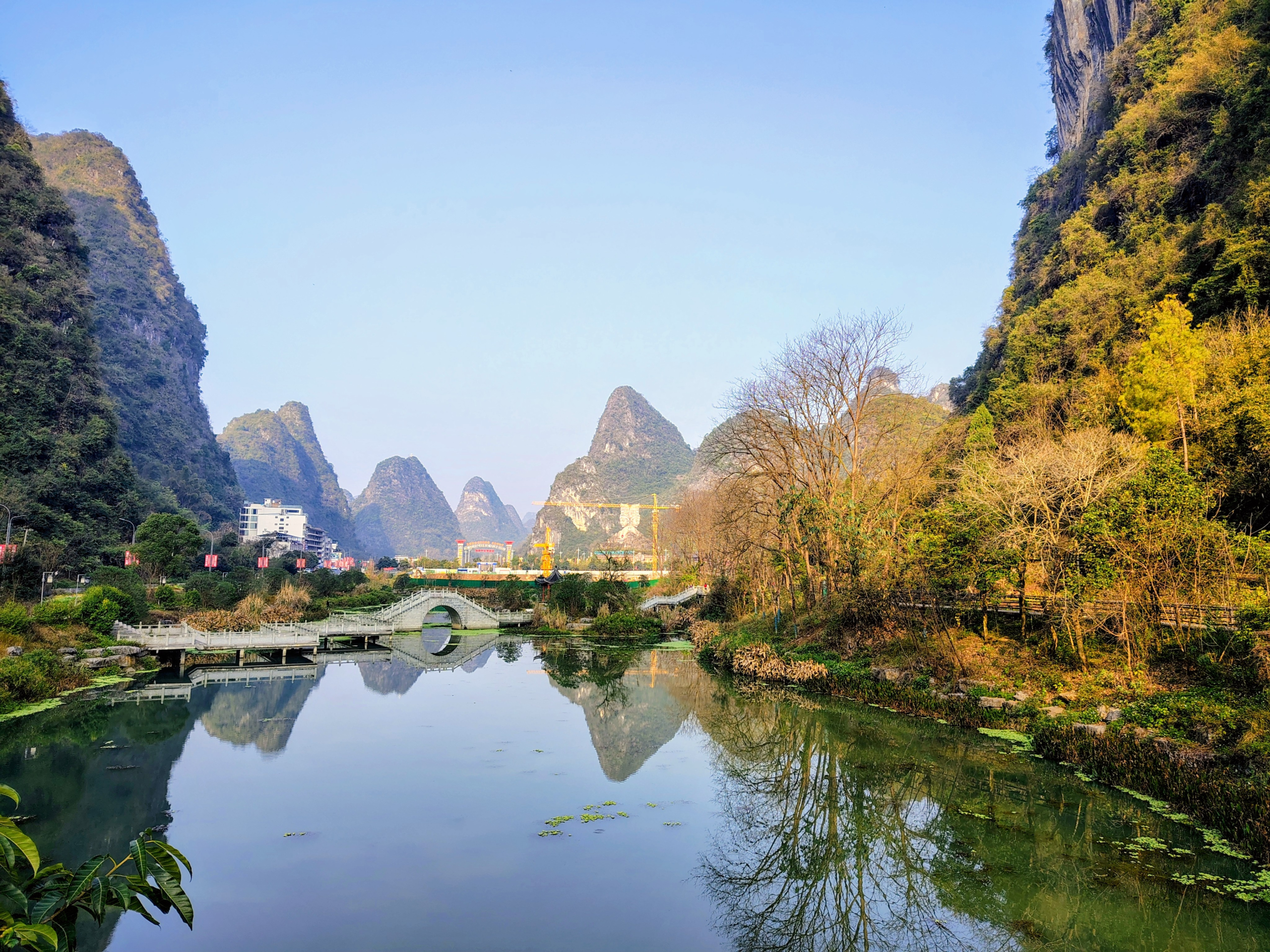A serene landscape of the Li River or a similar waterway in Yangshuo, China, featuring iconic limestone karst mountains reflected in the calm, green water. A traditional stone arched bridge crosses the water in the mid-ground, surrounded by lush trees and a clear blue sky. This scenic view highlights the natural beauty explored by a transgender man during his travels through China.