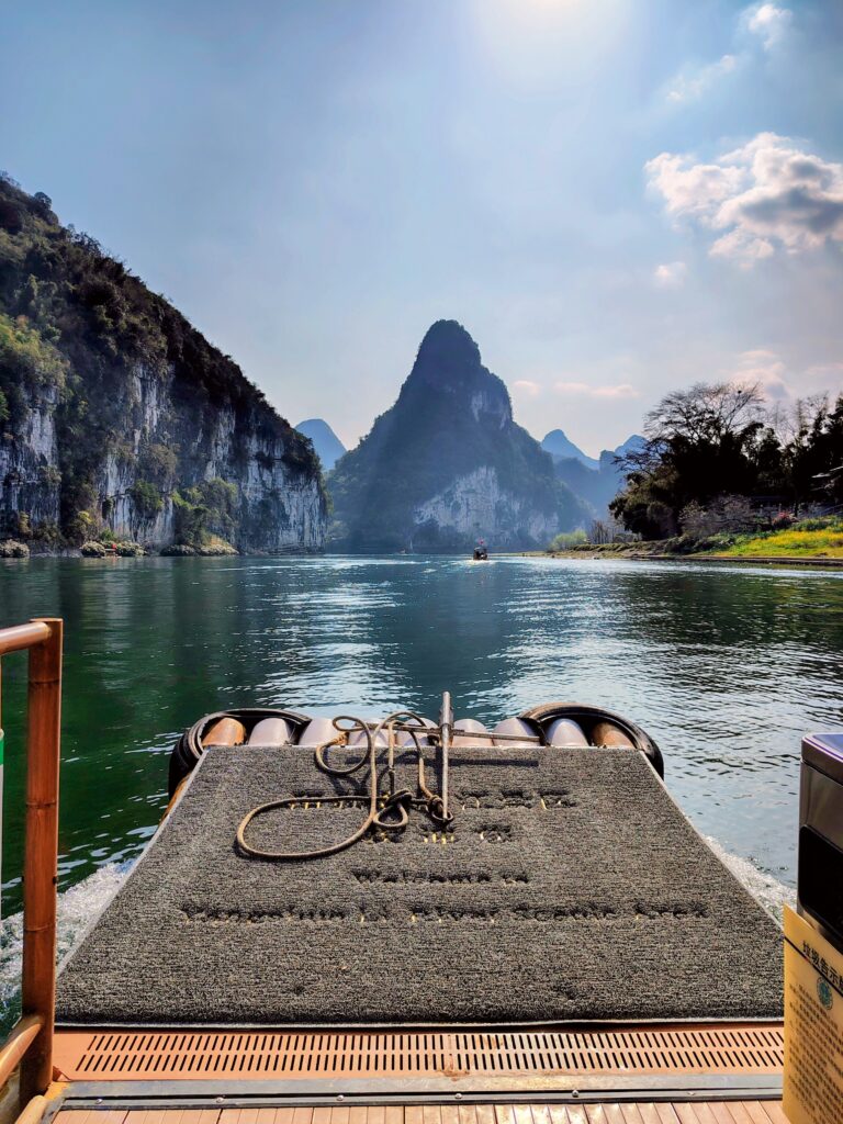 A first-person perspective from the front of a traditional bamboo raft cruising down the Li River in Yangshuo, China. A coiled rope sits on the raft's deck, leading the eye toward a towering, pointed karst mountain peak and lush green cliffs under a bright sun. This serene moment is a highlight of a trans masculine travel guide to Southern China.