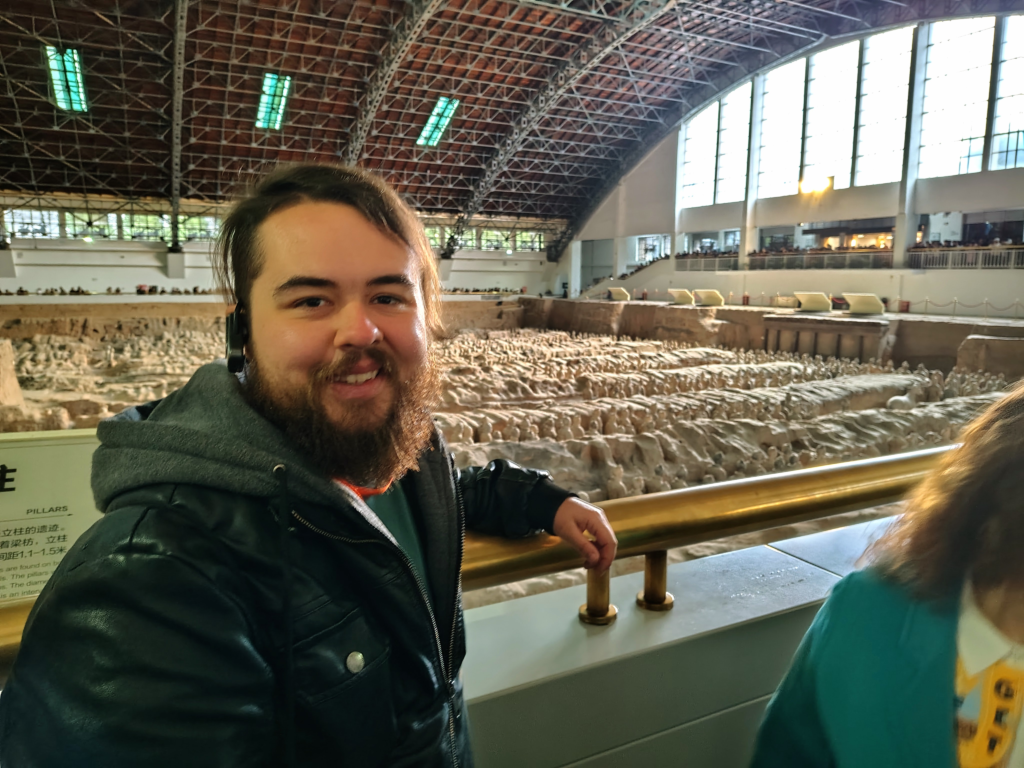 A smiling bearded transgender man stands at a railing overlooking the massive archaeological pit of the Terracotta Army in Xi'an, China. Thousands of life-sized clay soldier statues are visible in the background under a large, vaulted hangar roof. This photo captures a major bucket-list moment from an international travel blog focused on the trans masculine experience.